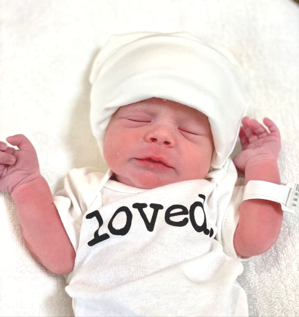 Newborn baby wearing a white onesie with 'loved' printed on it, and a seamless bamboo baby hat