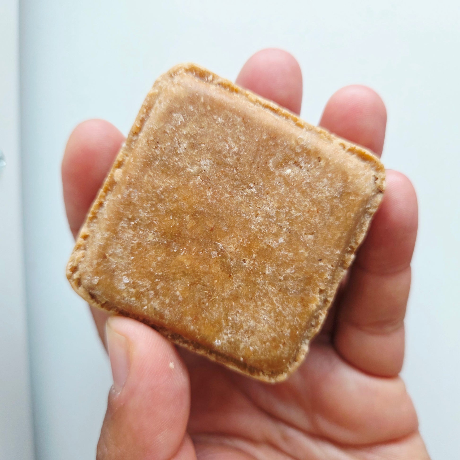 A hand holding a small, square, golden-Bare. Wash bar with a rich rooibos caramel colour and a textured surface.
