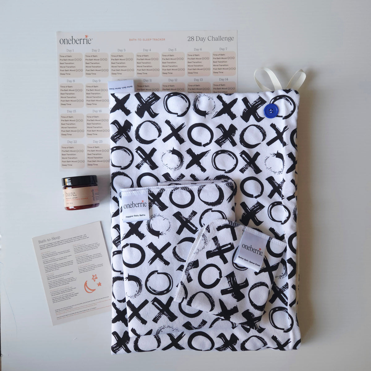 A Bath Bundle set by OneBerrie, featuring  Love you More XO black pattern on white background,, a matching bath swaddle, a washcloth, a 2oz Bare. Lotion, a 28-day challenge tracker sheet, and an instruction card on a light gray background