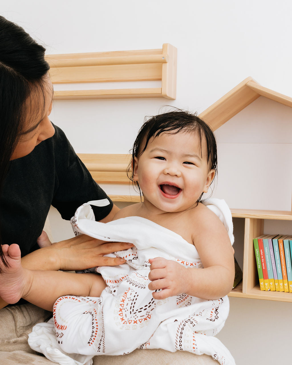 Little girl toddler wrapped in a Here comes the sun geometric rainbow towel laughing with mom holding her and smiling.