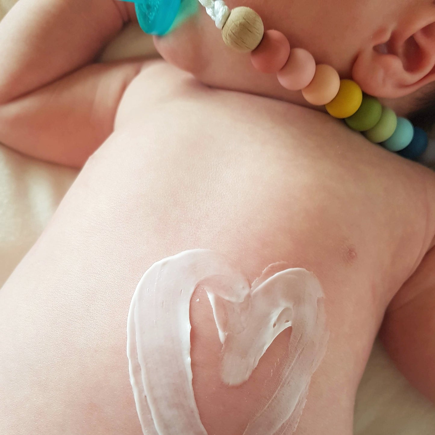 Close-up of BARE. Rooibos Lotion swirled in a heart shape on a newborn baby's back, with a rainbow silicone teether necklace visible
