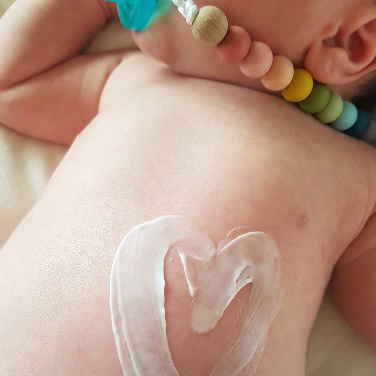 Close-up of BARE. Rooibos Lotion swirled in a heart shape on a newborn baby's back, with a rainbow silicone teether necklace visible