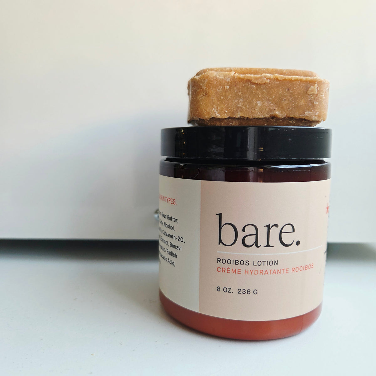 A jar of bare. Rooibos Lotion with a black lid and a light beige label, topped with a square of bare. wash syndet bar, on a white surface with a white background.