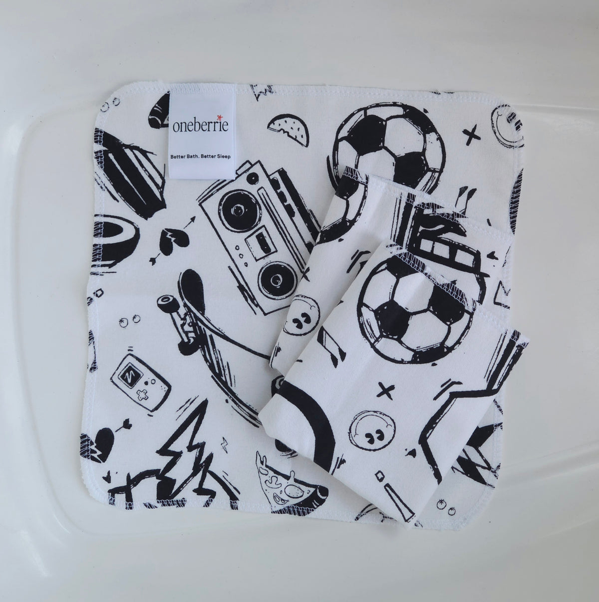 Three white flannel baby washcloths with a black graphic print of retro boomboxes, soccer balls, skateboards, and pizza slices, folded inside a clear plastic tub. The brand label 'oneberrie' is visible on the top left.