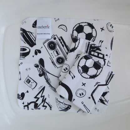 Three white flannel baby washcloths with a black graphic print of retro boomboxes, soccer balls, skateboards, and pizza slices, folded inside a clear plastic tub. The brand label 'oneberrie' is visible on the top left.