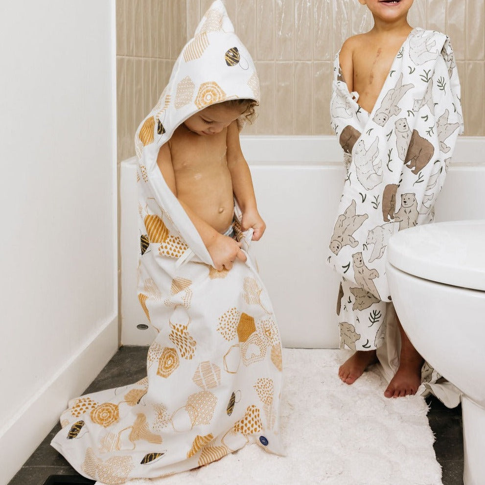 Two older free standing kids outside the bath wrapped in oneberrie hooded hands free towels. One girl is wearing the hood in the honeycomb bee print. the other is wrapped in a three bears print smiling. 