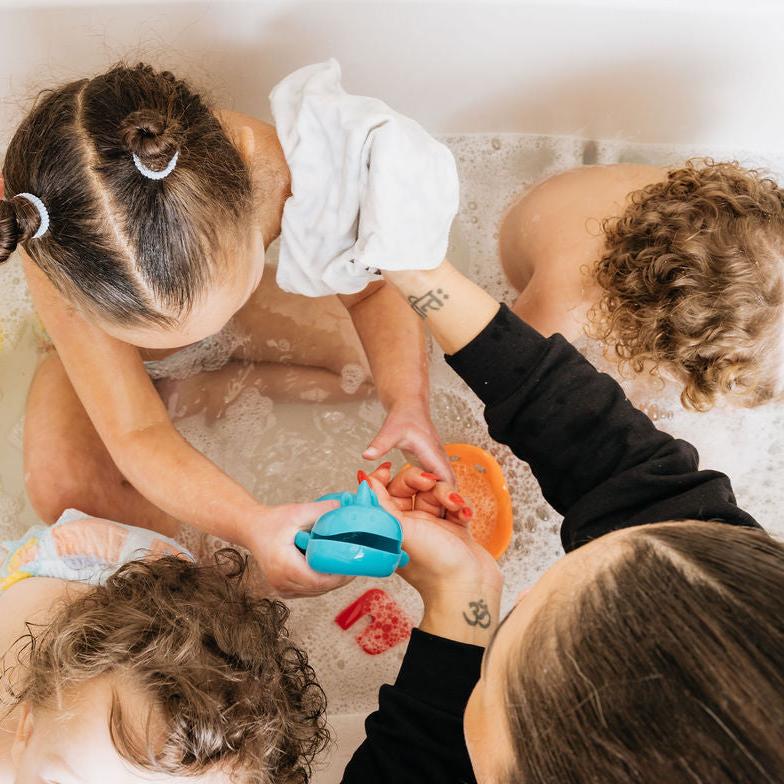 Mom washing three children, 2 with curly blonde hair and one with 2 twist buns, in the bath with bubbles and bath towels. Mom is using a Oneberrie washcloths
