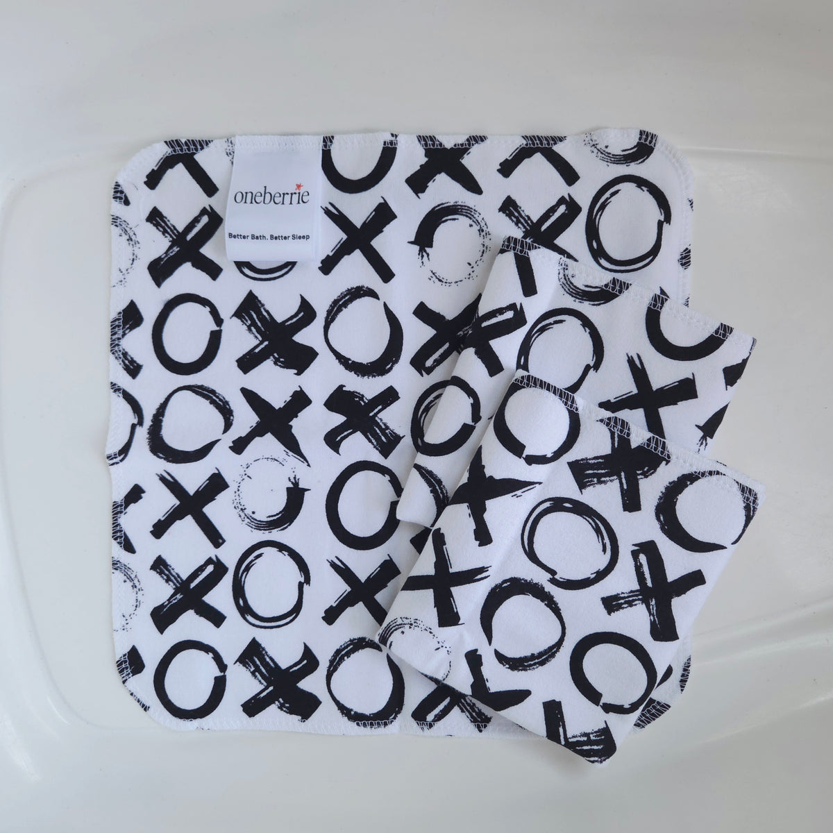 Three white flannel baby washcloths with a black graphic print of x's and o's, folded inside a clear plastic tub. The brand label 'oneberrie' is visible on the top left.