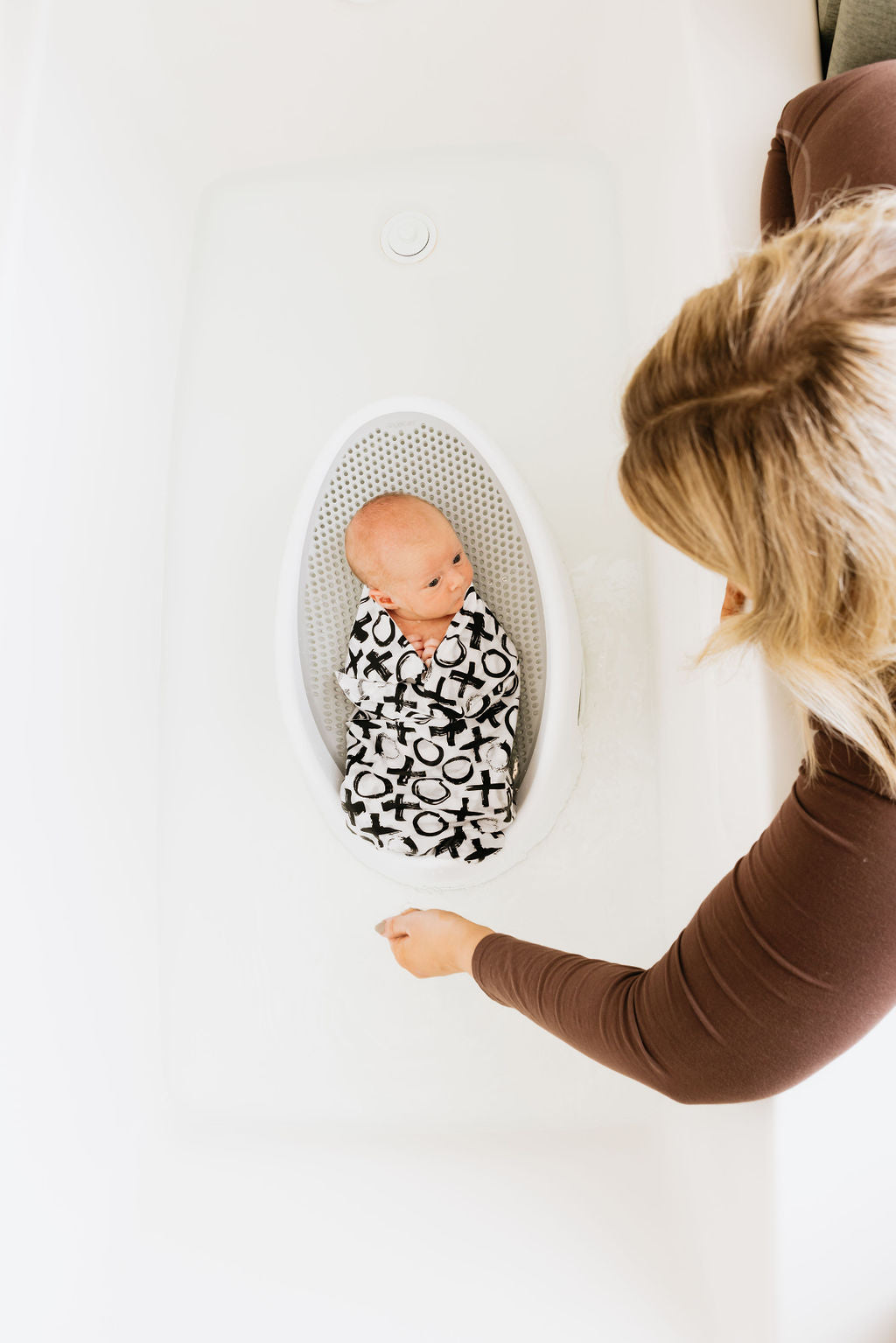 Newborn in a Mesh bath seat wrapped in a Oneberrie paint brush XO pattern with mom leaning over the tub 
