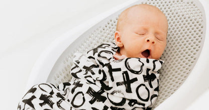 Newborn baby yawning wrapped in a XO swaddle in a plastic mesh bath seat.