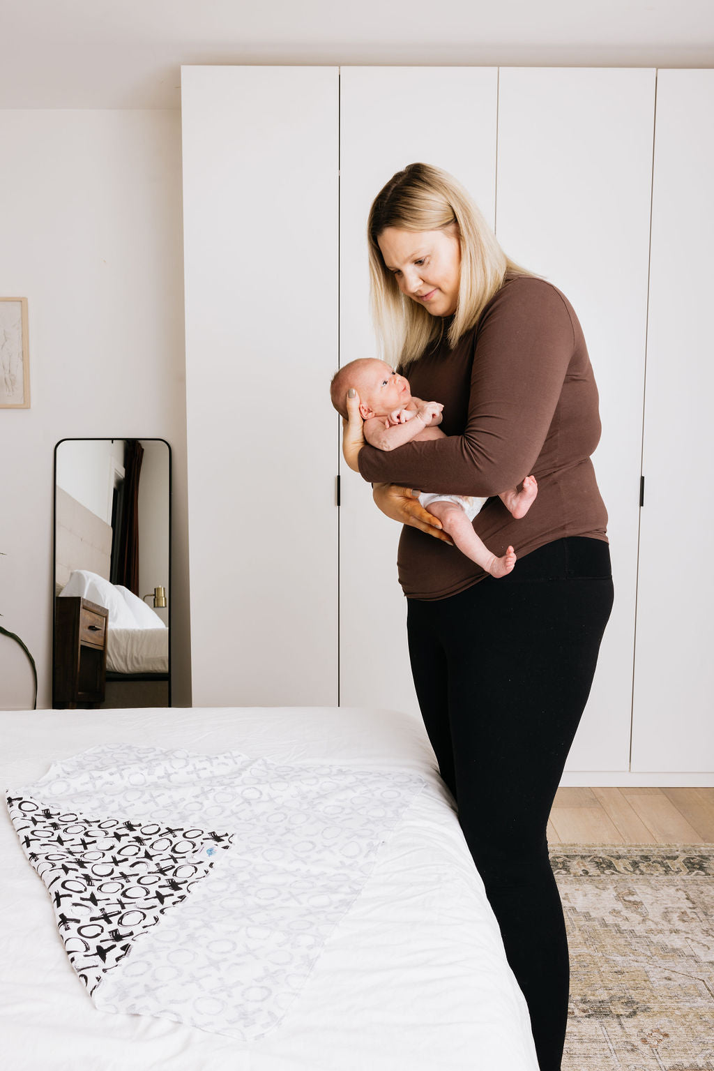Woman holding a baby in a bedroom with white walls and a bed.