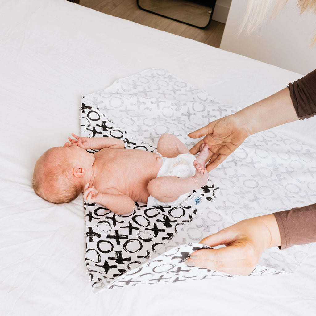Baby lying on a patterned blanket with a person adjusting it in a bedroom setting.