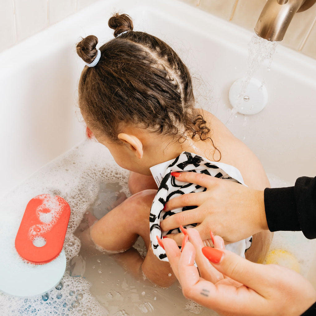 Child in a bathtub with bubbles, being bathed by an adult, surrounded by bath toys.
