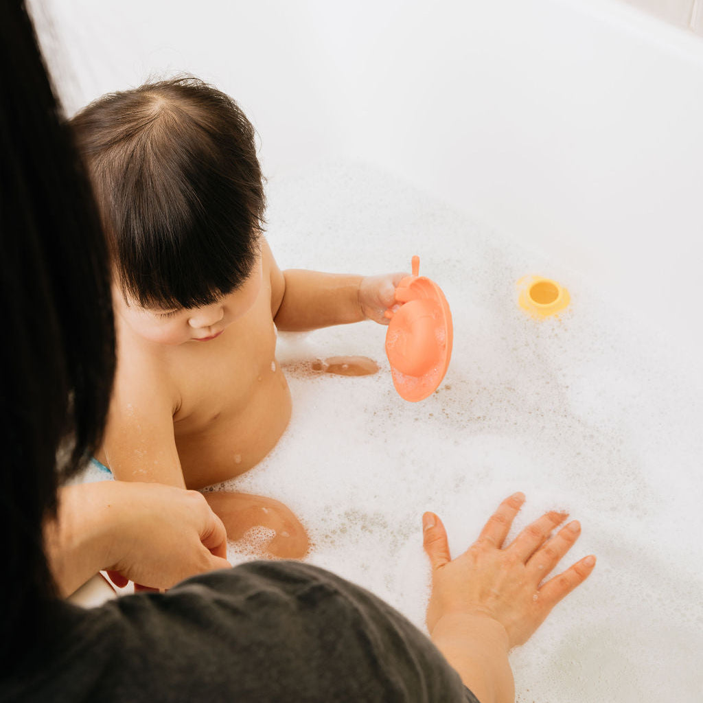 Child in a bathtub with bubbles, holding a toy, being bathed by an adult.