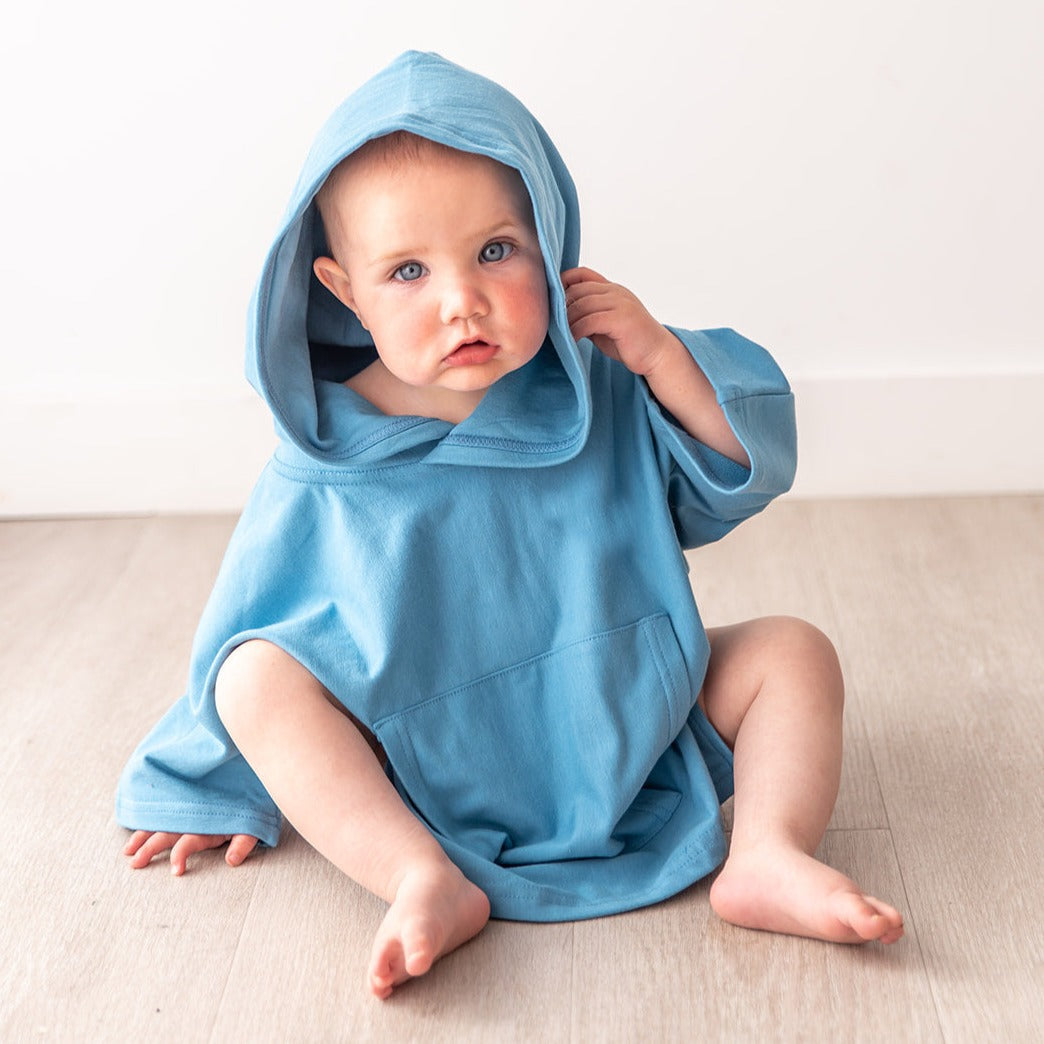 Blue eyed Baby wearing a blue hooded Oneberrie dolman Poncho sitting on a beige surface.