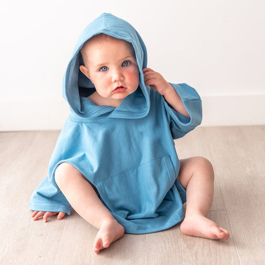 Blue eyed Baby wearing a blue hooded Oneberrie dolman Poncho sitting on a beige surface.