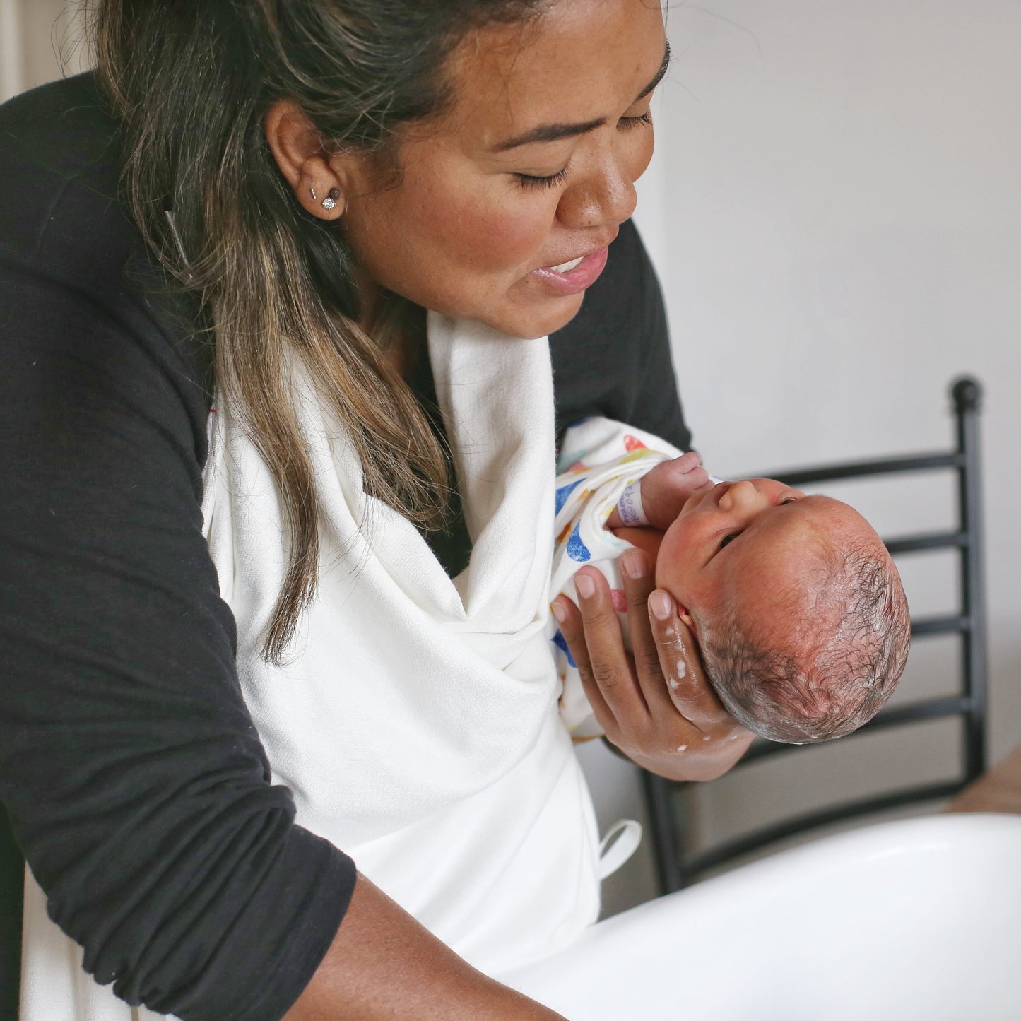 Woman holding a newborn and washing their hair in a football hold, wearing the towel and the baby is staring up at her