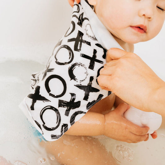 A baby wearing a white cotton bath towel with a black 'Tic-Tac-Toe' style print, held by an adult hand in a bubbly bathtub.