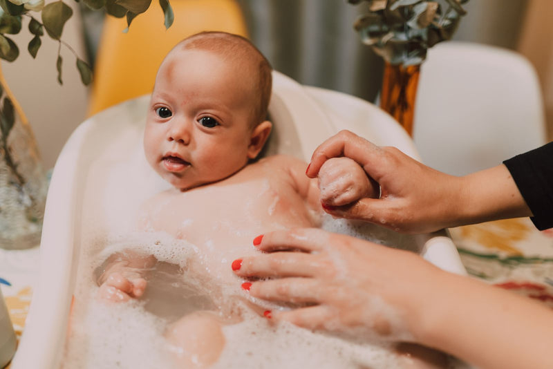 Calm baby sitting in a small tub of bubbly water while a parent gently washes them