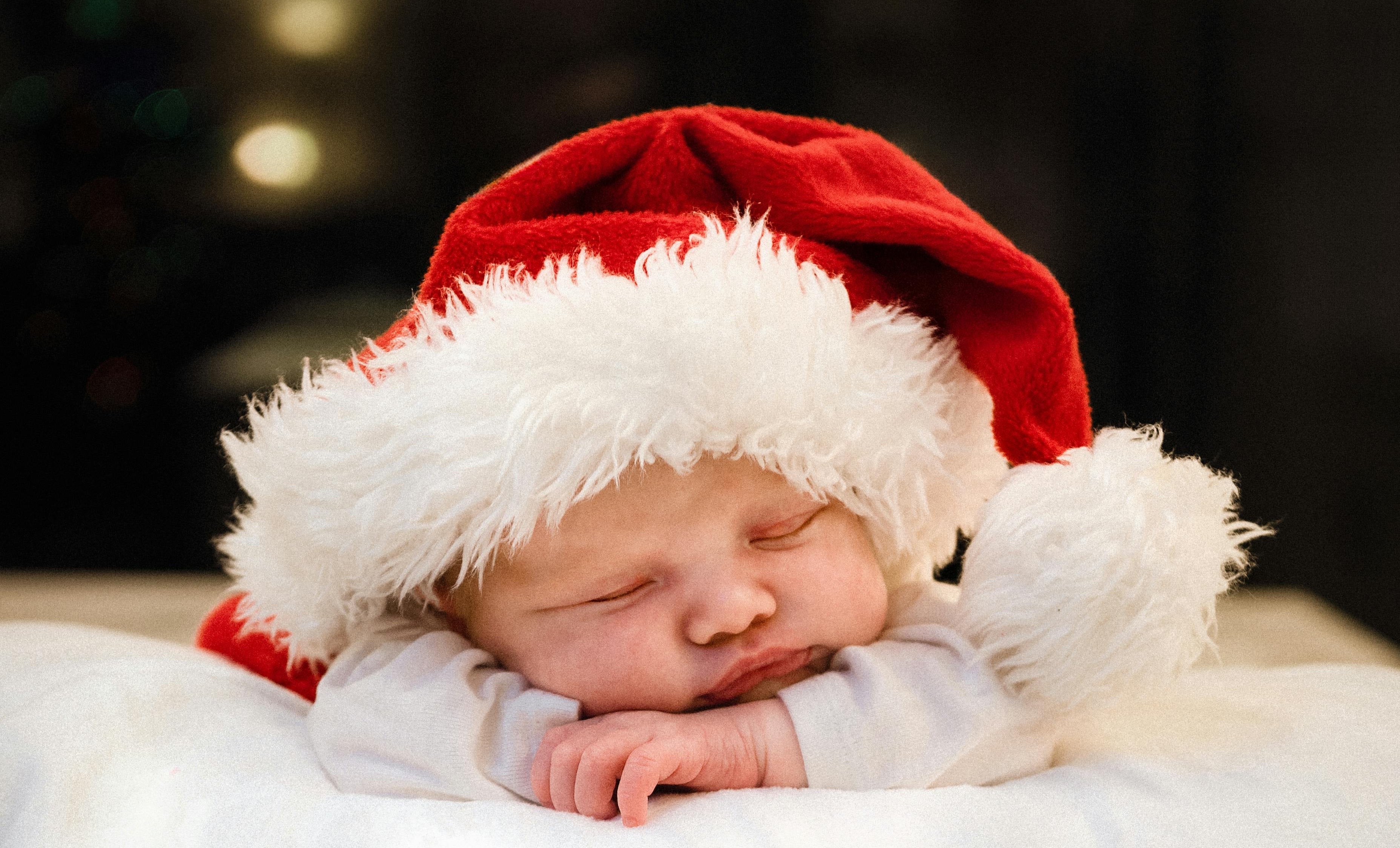 Newborn baby wearing a Santa hat with 'Babys 1st Christmas' text on a blanket.