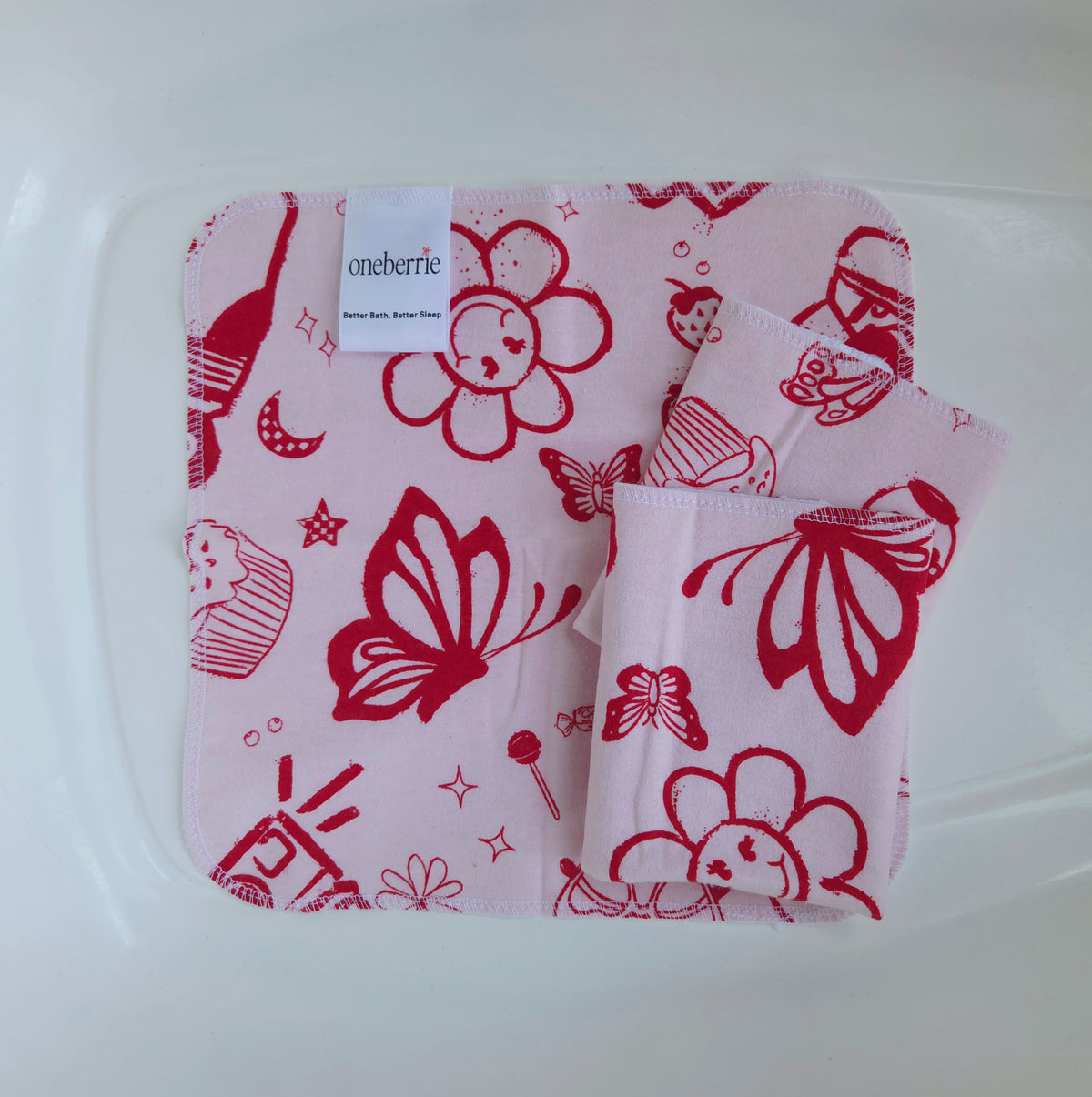 Three pink flannel baby washcloths with a red graphic print of retro flowers, butterfly, lollipop and cupcakes folded inside a plastic tub. The brand label 'oneberrie' is visible on the top left.