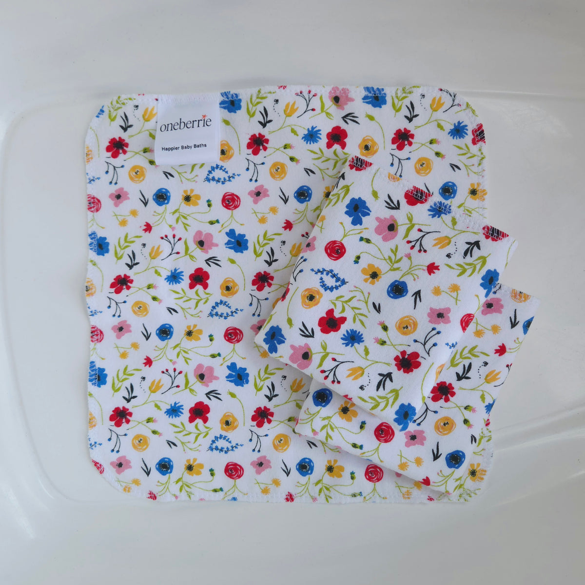 Three white flannel baby washcloths with a colorful wildflowers in pink, blue, yellow, red and greenery, folded inside a plastic tub. The brand label 'oneberrie' is visible on the top left.